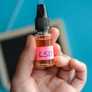 Liquid LSD, A small glass vial containing liquid LSD with a dropper cap, sitting on a surface. The vial is transparent, revealing a clear or slightly amber liquid inside. The dropper is positioned above the bottle, indicating the process of measuring out a precise dose.