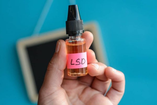 Liquid LSD, A small glass vial containing liquid LSD with a dropper cap, sitting on a surface. The vial is transparent, revealing a clear or slightly amber liquid inside. The dropper is positioned above the bottle, indicating the process of measuring out a precise dose.